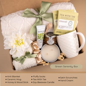 May include: A gift box with a white knit blanket, ceramic mug, honey, and a soy-beeswax candle. The box also contains tea, hand cream, and fluffy socks. The box is tied with a green ribbon and has the text "Green Serenity Box".
