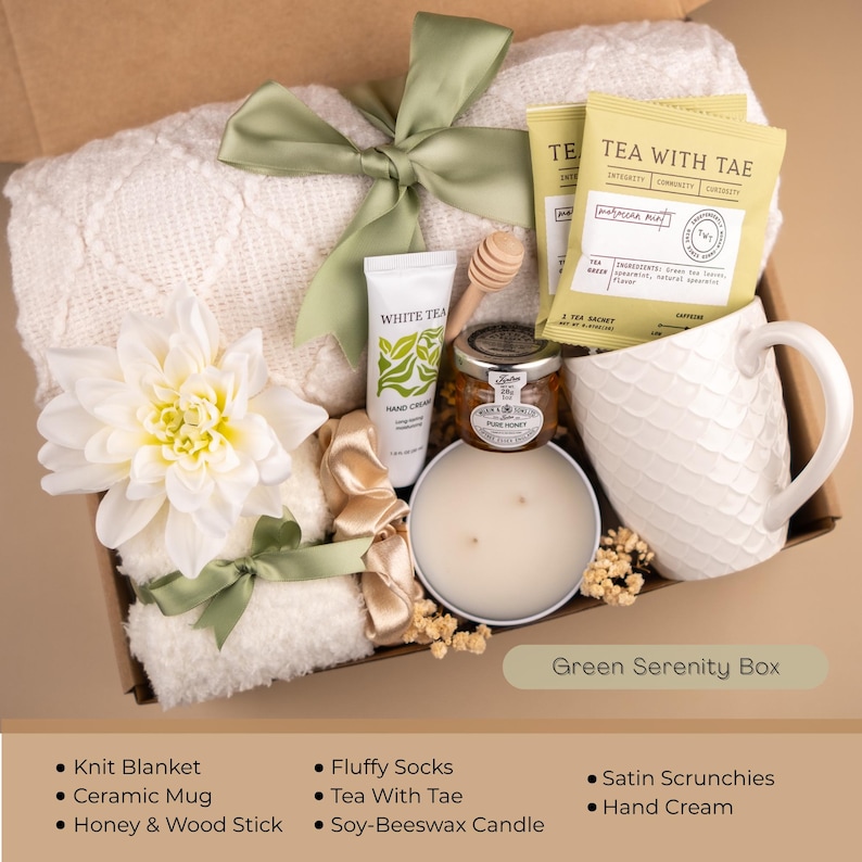 May include: A gift box with a white knit blanket, ceramic mug, honey, and a soy-beeswax candle. The box also contains tea, hand cream, and fluffy socks. The box is tied with a green ribbon and has the text "Green Serenity Box".