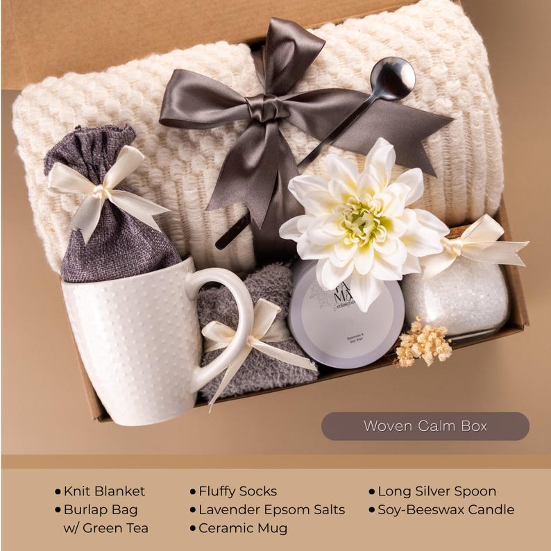 May include: A gift box filled with self-care items. It includes a white knit blanket, a ceramic mug, a burlap bag, fluffy socks, lavender Epsom salts, a soy-beeswax candle, and a long silver spoon. The box is tied with a gray ribbon and a white flower.