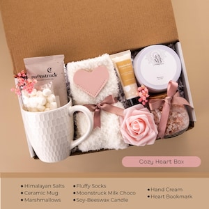 May include: A gift box labeled "Cozy Heart Box" contains a white ceramic mug filled with marshmallows, fluffy socks with a pink heart, hand cream, and a soy-beeswax candle. Other items include Himalayan salts, a heart bookmark, and Moonstruck milk chocolate.