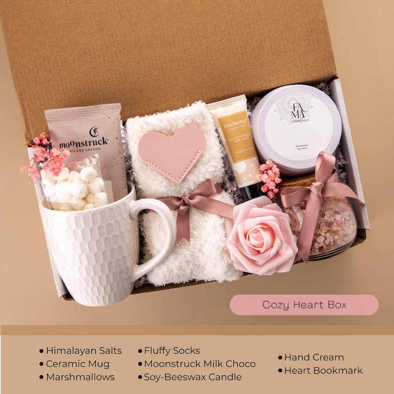 May include: A gift box labeled "Cozy Heart Box" contains a white ceramic mug filled with marshmallows, fluffy socks with a pink heart, hand cream, and a soy-beeswax candle. Other items include Himalayan salts, a heart bookmark, and Moonstruck milk chocolate.