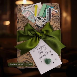 May include: A rectangular gift package wrapped in brown paper with a black floral rose pattern, tied with a wide olive green satin ribbon. Tucked inside are stickers, a yellow pen, and a white card with the text For fresh starts and healing stories and a small green heart-shaped stone.