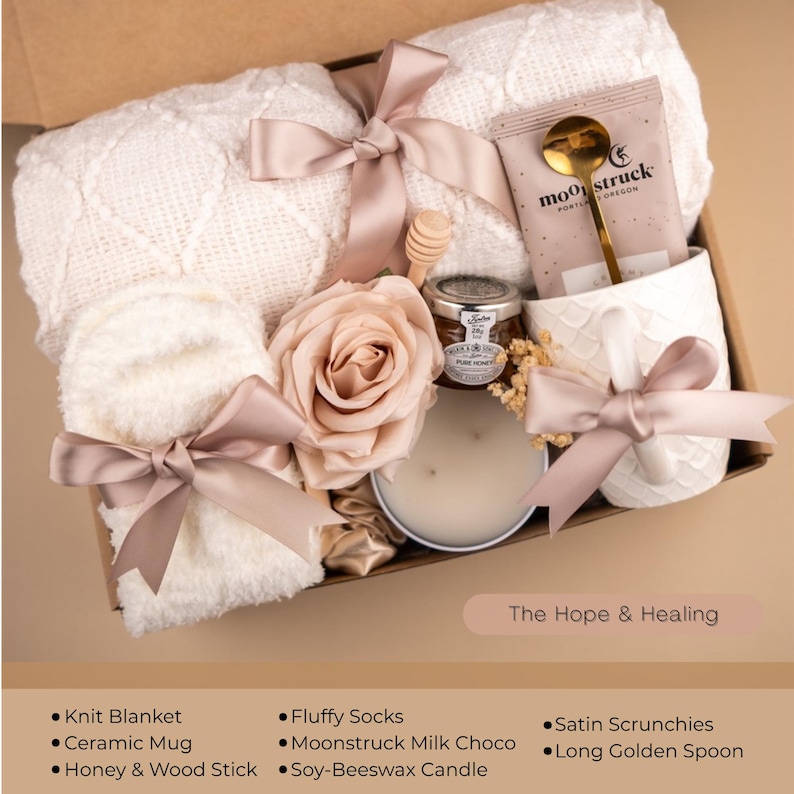 May include: A gift box filled with self-care items. It includes a white knit blanket, fluffy socks, a ceramic mug, a candle, a jar of honey, and a rose. The box is tied with a satin ribbon. The text on the box reads "The Hope & Healing".
