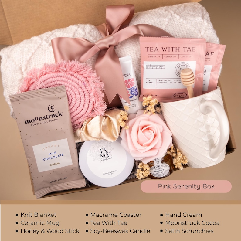 May include: A gift box filled with self-care items. It includes a white knit blanket, a pink macrame coaster, a ceramic mug, a hand cream, a Moonstruck cocoa packet, a honey and wood stick, a soy-beeswax candle, satin scrunchies, and tea packets. The box is tied with a pink ribbon.
