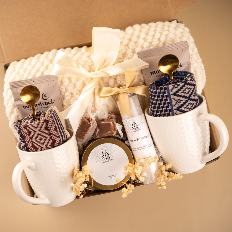 May include: A gift box filled with self-care items. Includes two white textured mugs with patterned socks, tea packets, a bottle of bath salts, a candle, and wrapped candies. A cream-colored blanket and ribbon complete the presentation.