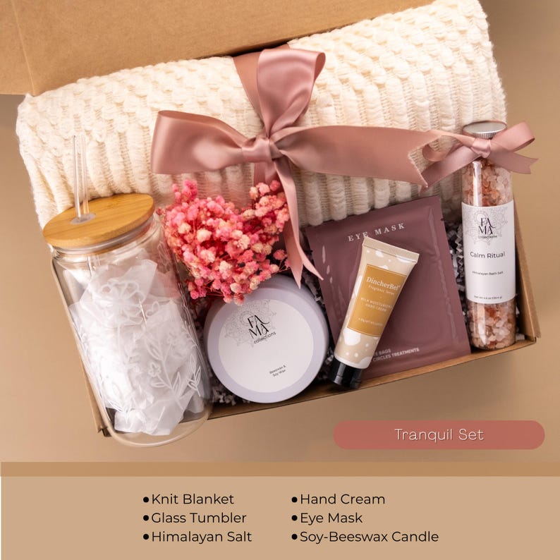 May include: A gift box with a cream-colored knit blanket, glass tumbler, pink flowers, eye mask, hand cream, Himalayan salt, and a soy-beeswax candle. The box is tied with a pink ribbon and labeled "Tranquil Set."