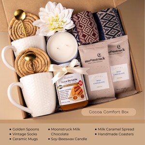 May include: A gift box filled with comfort items. It includes white ceramic mugs, golden spoons, a soy-beeswax candle, Moonstruck milk chocolate, a jar of dulce de leche, patterned socks, and handmade coasters. The box is labeled "Cocoa Comfort Box."