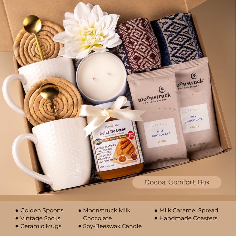 May include: A gift box filled with comfort items. It includes white ceramic mugs, golden spoons, a soy-beeswax candle, Moonstruck milk chocolate, a jar of dulce de leche, patterned socks, and handmade coasters. The box is labeled "Cocoa Comfort Box."