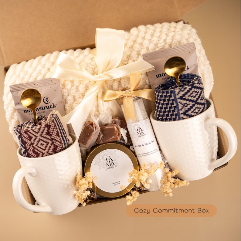 Peut inclure: Un coffret cadeau contenant une couverture cr&egrave;me, deux tasses blanches textur&eacute;es avec des chaussettes &agrave; motifs, des sachets de th&eacute;, une cuill&egrave;re dor&eacute;e, un pot, une bouteille et des bonbons emball&eacute;s. Le coffret est nou&eacute; avec un ruban cr&egrave;me. Le texte sur le coffret indique "Cozy Commitment Box."