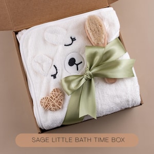 May include: A white hooded baby towel with bear face embroidery, a wooden brush, and a woven heart are presented in a cardboard box. A sage green ribbon is tied around the towel. The text "SAGE LITTLE BATH TIME BOX" is visible.