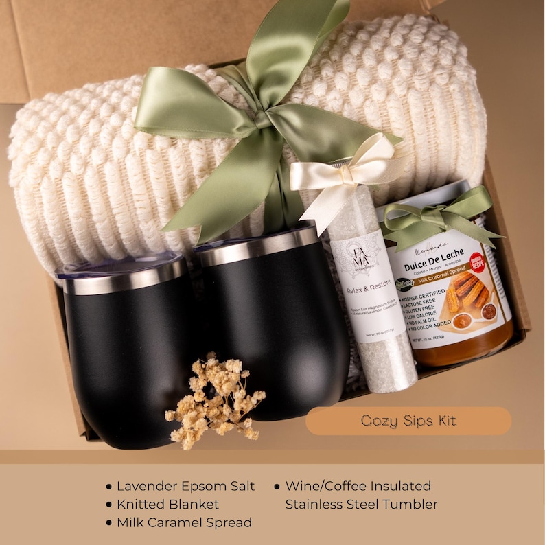 May include: A gift box with a cream-colored knitted blanket, two black stainless steel tumblers, a jar of Dulce De Leche, and a bottle of lavender Epsom salt. The box is tied with a green ribbon and includes the text "Cozy Sips Kit".