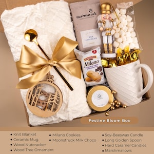 Golden holiday gift box with knit blanket, candle, ceramic mug, nutcracker, ornament, and cocoa — luxury Christmas care package for women or cozy winter gift.