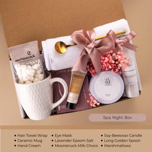 May include: A spa night gift box with a white ceramic mug, marshmallows, a hair towel wrap, and a golden spoon. The box includes a soy-beeswax candle, hand cream, and lavender Epsom salt. The box is tied with a pink ribbon.