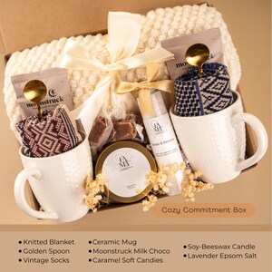 May include: A gift box filled with a cream-colored knitted blanket, two white ceramic mugs, and various self-care items. Includes socks, a candle, bath salts, chocolates, and golden spoons. The box is tied with a cream ribbon and labeled "Cozy Commitment Box."