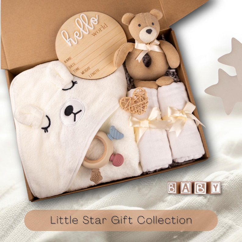May include: A baby gift set in a cardboard box. The set includes a teddy bear, a hooded towel with a bear face, a wooden sign that says "hello world", a wooden heart, a teether, and two rolled white towels tied with a ribbon. The text "Little Star Gift Collection" is at the bottom.