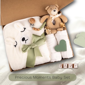 May include: A baby gift set in a cardboard box, featuring a white llama-shaped blanket, a teddy bear rattle, and a pacifier clip. The set includes white washcloths with a green heart and a green ribbon. The text reads "Precious Moments Baby Set".