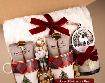 May include: A holiday gift box with a white towel, two mugs with Christmas tree designs, and a candle. The mugs have red plaid bands and gold spoons. A wooden ornament and nutcracker are also included. The box is labeled "Holiday Season Gift Box" and "Luxe Christmas Box."