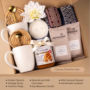 Peut inclure: Un coffret cadeau rempli d'articles de confort. Il comprend des tasses en c&eacute;ramique blanches, des cuill&egrave;res dor&eacute;es, une bougie en cire de soja, du chocolat au lait Moonstruck, du Dulce De Leche, des chaussettes vintage et des dessous de verre faits &agrave; la main. Le coffret est &eacute;tiquet&eacute; "Cocoa Comfort Box".