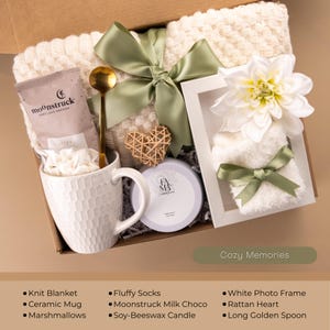 May include: A gift box filled with a knit blanket, ceramic mug, marshmallows, fluffy socks, Moonstruck milk chocolate, soy-beeswax candle, white photo frame, rattan heart, and a long golden spoon. The box is tied with a green ribbon and has the text "Cozy Memories".