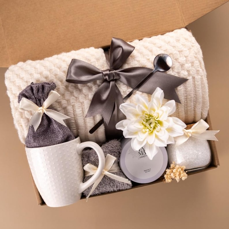 May include: A gift box containing a cream-colored textured throw blanket tied with a dark gray ribbon, a white ceramic mug with a dotted pattern, a gray fabric pouch, a gray candle, a small glass jar with white contents, and a white flower.