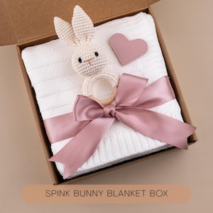 May include: A white baby blanket with a knitted texture, a crochet bunny rattle, and a pink heart-shaped tag, all presented in a cardboard box. A pink satin ribbon is tied around the blanket. The text "SPINK BUNNY BLANKET BOX" is visible.