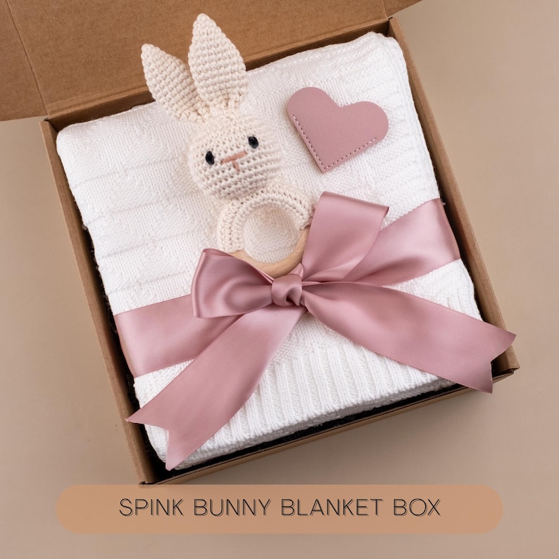 May include: A white baby blanket with a knitted texture, a crochet bunny rattle, and a pink heart-shaped tag, all presented in a cardboard box. A pink satin ribbon is tied around the blanket. The text "SPINK BUNNY BLANKET BOX" is visible.