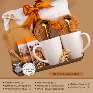 Peut inclure: Un coffret cadeau avec une serviette de cuisine blanche, deux tasses en c&eacute;ramique blanches, une planche &agrave; d&eacute;couper en bambou et d'autres articles. Le coffret comprend des sachets de noix m&eacute;lang&eacute;es, un pot de miel, un b&acirc;tonnet en bois, des fleurs s&eacute;ch&eacute;es, des dessous de verre en rotin et des cuill&egrave;res dor&eacute;es.