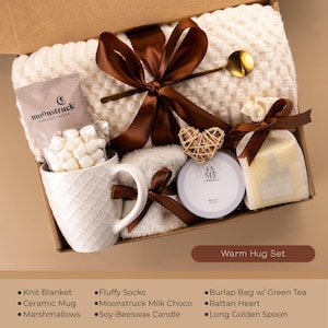 May include: A gift box labeled "Warm Hug Set" contains a cream-colored knit blanket, ceramic mug with marshmallows, a soy-beeswax candle, and a burlap bag with green tea. The box is tied with a brown ribbon and includes a golden spoon and a rattan heart.