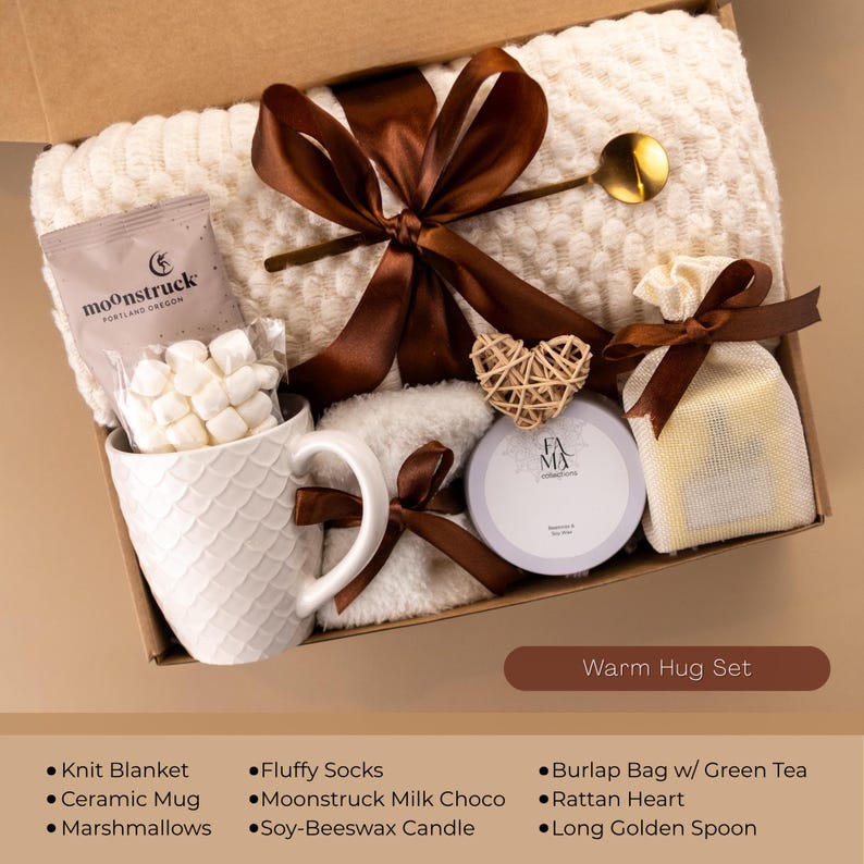 May include: A gift box labeled "Warm Hug Set" contains a cream-colored knit blanket, ceramic mug with marshmallows, a soy-beeswax candle, and a burlap bag with green tea. The box is tied with a brown ribbon and includes a golden spoon and a rattan heart.
