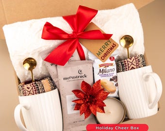May include: A holiday gift box filled with a white towel, two white mugs with patterned sleeves, gold spoons, chocolate, a candle, and a red bow. The box includes a tag that says "Merry Christmas". The text "Holiday Cheer Box" is also visible.