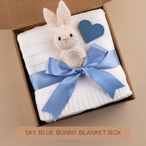 May include: A gift box containing a white baby blanket, a crochet bunny rattle, and a blue heart-shaped tag. The blanket is tied with a sky blue satin ribbon. The box has the text "SKY BLUE BUNNY BLANKET BOX".