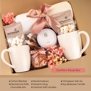 May include: A gift box filled with self-care items. Includes a white cotton blanket, two white ceramic mugs, marshmallows, a jar of caramel candies, a Himalayan salt jar, and a soy-beeswax candle. The box is tied with a pink ribbon. Text on the box reads "Comfort Ritual Box."