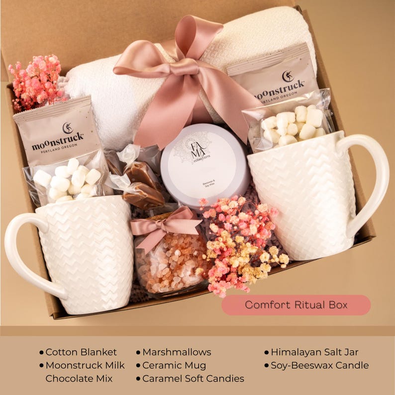 May include: A gift box filled with self-care items. Includes a white cotton blanket, two white ceramic mugs, marshmallows, a jar of caramel candies, a Himalayan salt jar, and a soy-beeswax candle. The box is tied with a pink ribbon. Text on the box reads "Comfort Ritual Box."