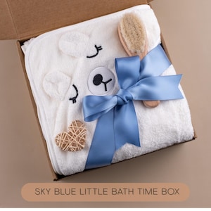 May include: A white hooded baby towel with a bear face design, a wooden brush, and a woven heart-shaped decoration, all presented in a cardboard box. A sky blue ribbon is tied around the towel. The text "SKY BLUE LITTLE BATH TIME BOX" is at the bottom.