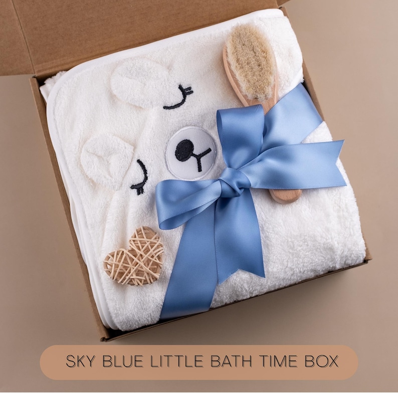 May include: A white hooded baby towel with a bear face design, a wooden brush, and a woven heart-shaped decoration, all presented in a cardboard box. A sky blue ribbon is tied around the towel. The text "SKY BLUE LITTLE BATH TIME BOX" is at the bottom.