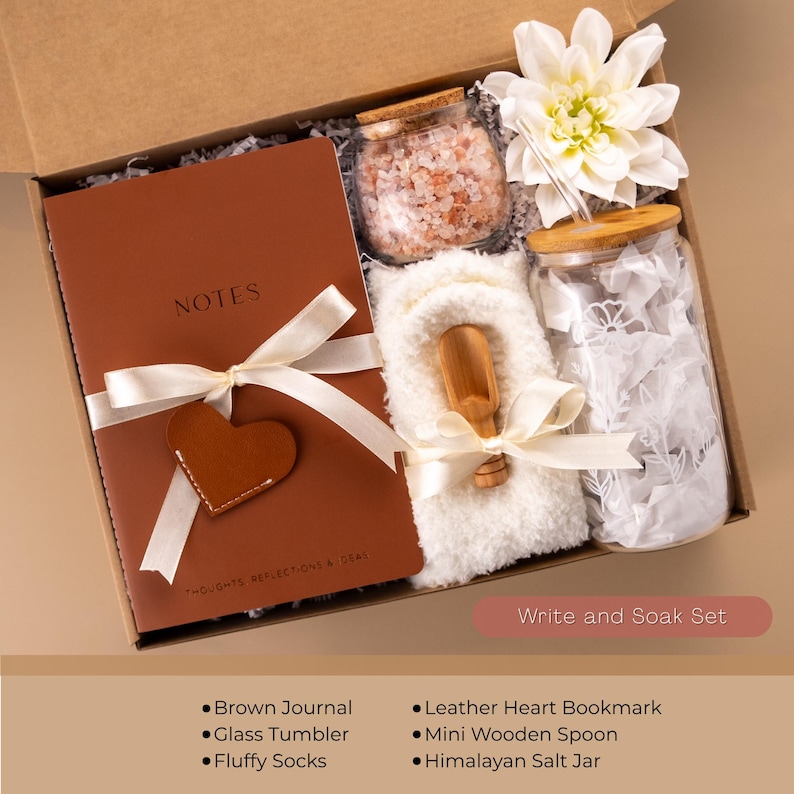 May include: A gift box labeled "Write and Soak Set" contains a brown journal, glass tumbler, fluffy socks, leather heart bookmark, mini wooden spoon, and Himalayan salt jar. The journal is tied with a white ribbon and has a leather heart. A white flower is also included.