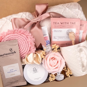 May include: A gift box containing a white textured blanket with a dusty pink ribbon, two Tea With Tae sachets, a wooden honey dipper, a white mug, a Moonstruck milk chocolate bar, a pink fringe coaster, a gold scrunchie, a candle, and a pink rose.