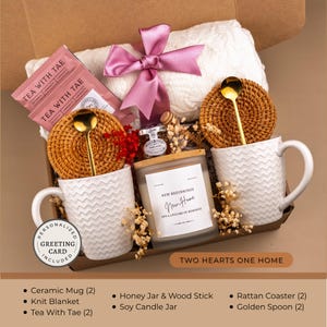 Peut inclure: Un coffret cadeau avec une couverture en tricot blanche, deux tasses en c&eacute;ramique blanches, des sachets de th&eacute;, une bougie de soja, un pot de miel, des dessous de verre en rotin et des cuill&egrave;res dor&eacute;es. La bougie indique "New Beginnings, New Home, and a lifetime of memories."
