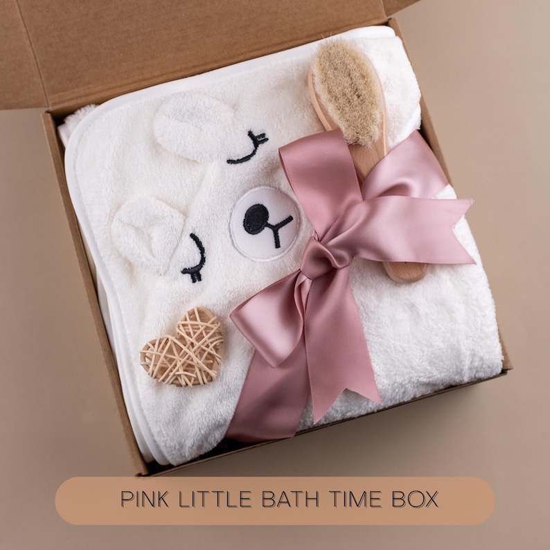 May include: A white hooded baby towel with a bear face design, a wooden brush, and a woven heart-shaped decoration are presented in a cardboard box. A pink satin ribbon is tied around the towel. The box has the text "PINK LITTLE BATH TIME BOX".
