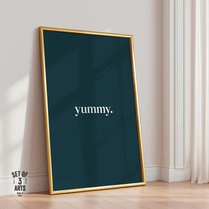 Könnte beinhalten: Ein gerahmter Kunstdruck mit einem goldfarbenen Rahmen. Das Kunstwerk zeigt einen dunkelblauen Hintergrund mit dem Wort "yummy" in Weiß, zentriert. Der Druck lehnt an einer weißen Wand und einem Holzboden.