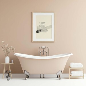 Op de afbeelding: Een ingelijste print met de tekst "1. LIFT. 2. AIM. 3. FLUSH. 4. REPEAT." hangt boven een badkuip met klauwpoten. De badkamer heeft een neutraal kleurenpalet met een kleine houten bijzettafel en een handdoekenrek.