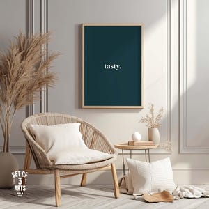 Könnte beinhalten: Ein gerahmter tealfarbener Druck mit dem Wort "tasty" in weißen Buchstaben hängt über einem geflochtenen Stuhl mit einem weißen Kissen. Ein kleiner Beistelltisch und dekorative Kissen vervollständigen das gemütliche Innendesign. Getrocknetes Pampasgras sorgt für einen natürlichen Touch.