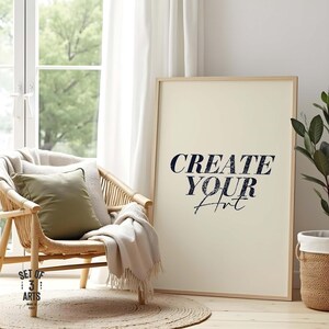 Può includere: Una stampa artistica incorniciata con la scritta "CREATE YOUR Art" in blu navy. L'opera d'arte è appoggiata a una cornice in legno chiaro. Una sedia in vimini con un cuscino verde e una coperta beige è in primo piano. Sono visibili anche un tappeto in juta e una pianta in vaso.