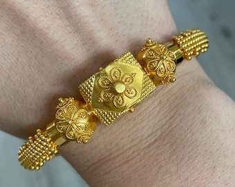 May include: A gold-colored bracelet with intricate floral and geometric designs. The bracelet features a central square element with a flower design and smaller floral accents on the sides. The ends of the bracelet have a beaded texture.