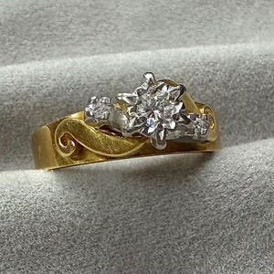 May include: A gold ring featuring a central cluster of small, clear, faceted stones set in a white metal mount. The band has a smooth, polished gold finish with decorative, swirling, raised gold accents on either side of the stone setting.