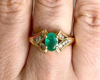 May include: A gold ring featuring an oval-cut emerald center stone. The ring is accented with small diamonds and has a decorative, openwork design on the sides. The ring is set on a gold band.