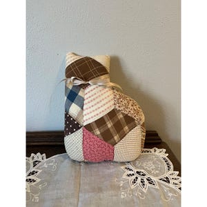 May include: A patchwork cat-shaped decorative item. The cat is made of various fabric patterns, including brown plaid, floral, and polka dots. A beige ribbon is tied around the neck. The item is sitting on a table with a white lace doily.