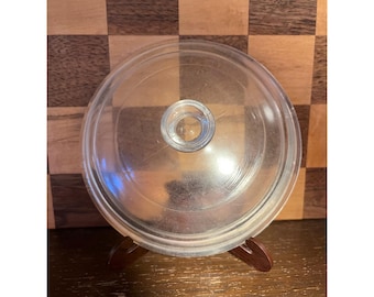 Könnte beinhalten: Ein klarer Glasdeckel mit einem zentralen Knopf, wahrscheinlich für einen Topf oder eine Pfanne. Der runde Deckel liegt auf einem kleinen, dunklen Holzständer. Der Hintergrund zeigt ein Holzschachbrettmuster.