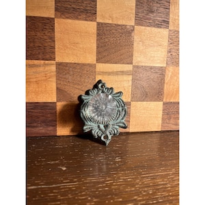 May include: An antique, ornate metal drawer pull with a clear, faceted glass center. The metal has a distressed, verdigris finish. The drawer pull is set against a wooden background with a checkerboard pattern.