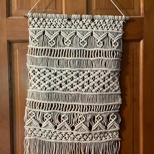 May include: A cream-colored macrame wall hanging with intricate knotting patterns and a fringed bottom. The wall decor is suspended from a wooden dowel and a white cord. The design features geometric shapes and vertical lines, creating a textured, bohemian aesthetic.